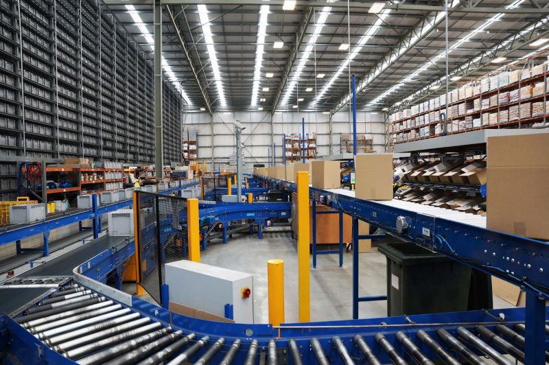 A wide, interior shot showcases a brightly lit warehouse or distribution centre, equipped with an extensive network of blue conveyor belt systems manufactured by Dyno Conveyors (dyno.co.nz). Various cardboard boxes are positioned on different sections of the conveyors, suggesting an active process of sorting or transportation. High shelving units filled with goods line the walls in the background, indicating a large-scale storage operation. The metal ceiling structure is visible overhead, supported by numerous pillars and illuminated by rows of artificial lighting.